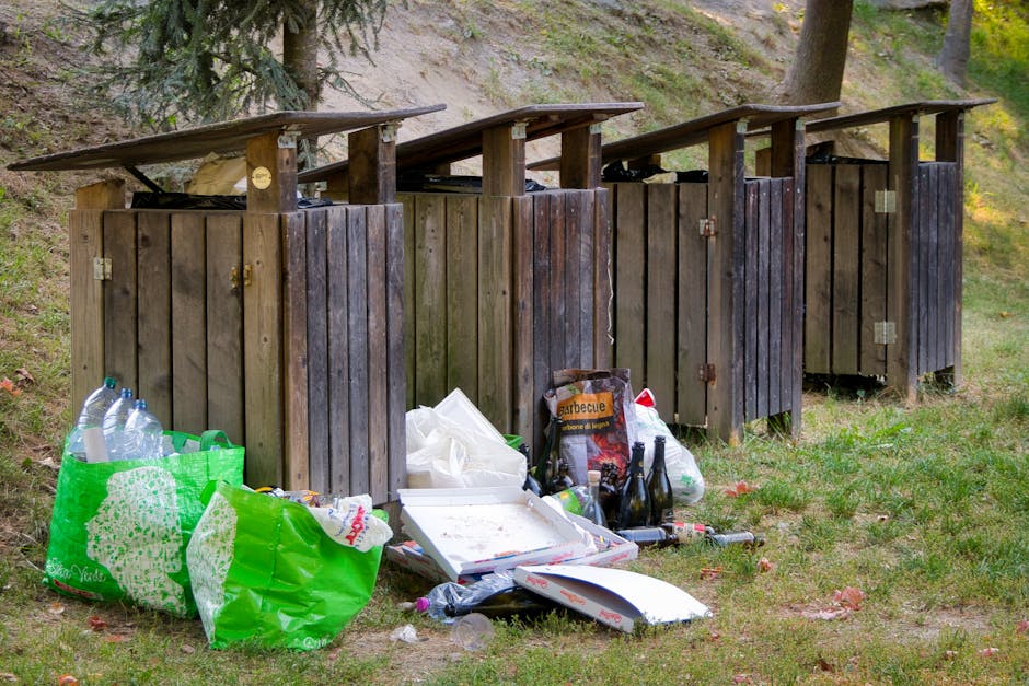 The image depicts a row of four outdoor wooden rubbish bins with slanted, weathered dark brown lids situated on a grassy area adjacent to a slight incline with trees and shrubs in the background. The bins are made of vertical wooden planks with metallic hinges and locks visible on some of the doors. In front of the bins, there is a collection of discarded waste including empty plastic bottles, some still in the large green recyclable shopping bag, a cardboard pizza box, a bag of charcoal labeled 'Barbecue', and various other miscellaneous items such as paper and plastic packaging. The waste appears to be loosely arranged on the ground, with some items leaning against the bins and others scattered around. The environment shows natural daylight, highlighting the textures of the wooden structures and the vibrant colors of the waste materials, which may indicate an instance of improper waste disposal outside designated rubbish collection points. This scene reflects a situation where private rubbish handling, such as individual or informal collection, is relevant, emphasizing the importance of accessible waste management solutions for residential outdoor areas.