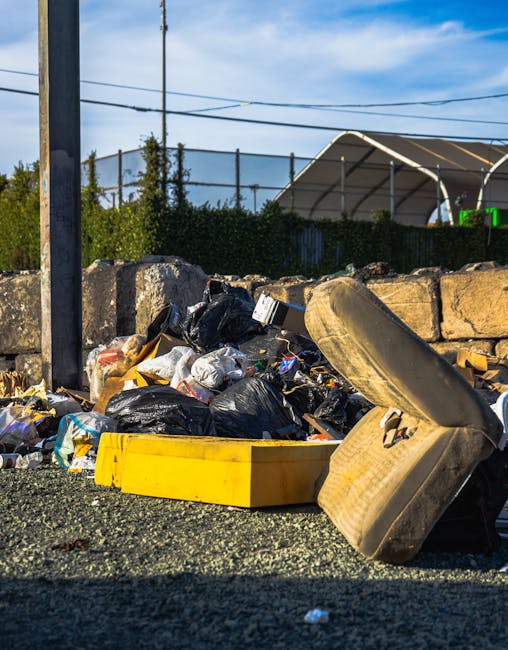 A sanitation worker wearing a yellow high-visibility vest and black trousers is actively loading waste into the rear compactor of a large red garbage truck parked at the edge of a paved street. The truck's back door is open, revealing the loading mechanism and the interior chamber where waste is compacted, with visible hydraulic components and metal surfaces. The worker is holding a large black garbage bag, which is partially inside the truck, and is surrounded by scattered rubbish on the ground, including pieces of paper and plastic. The scene takes place outdoors on a cloudy day, with some greenery, trees, and utility poles in the background, as well as a hint of nearby buildings and a blue signpost. The image illustrates typical urban waste collection activity, aligning with private rubbish removal services that handle on-site disposal and collection outside of municipal systems, fitting within the context of alternative waste management methods.
