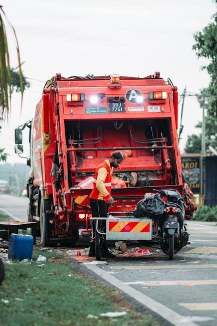 A waste collection worker operating a large red garbage truck on a street, with the truck's rear hatch open as the worker sorts through collected rubbish. The worker, dressed in a yellow high-visibility vest over a red shirt, is standing next to a metal barrier and a small black motorcycle parked adjacent to the truck. The scene takes place on a paved road with a painted curb, and there are utility poles, trees, and a building or shop in the background. The rubbish includes black plastic bags and other debris, some of which has spilled onto the ground nearby. The environment appears to be an urban or suburban street, with soft natural lighting indicating daytime. This image may relate to private waste collection services, such as those offered by rubbish removal companies, highlighting stages of on-site clearance or waste handling outside of municipal rubbish collection. Occasionally, the scene reflects the importance of proper rubbish disposal and the potential alternatives to traditional municipal services, as seen in independent waste removal operations.