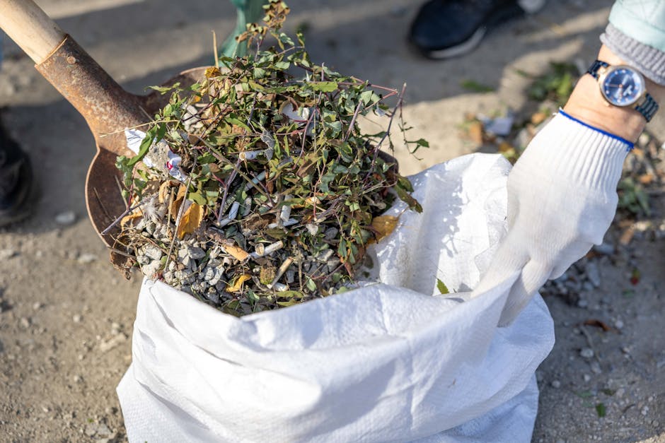 A person wearing a white work glove is holding a large white bag filled with garden debris, including small branches, leaves, and gravel. The bag appears to be made of woven plastic material and is open at the top, showing the contents clearly. A metal shovel with a rusted blade is resting against the bag, partially filled with similar garden waste. In the background, a gravel or dirt surface is visible, with some scattered leaves and debris. The person is wearing a light-colored long sleeve shirt or sweater with a blue watch on their wrist. The scene is outdoors, illuminated by natural daylight, suggesting an on-site garden clearance or waste collection activity, which aligns with private or alternative waste handling services often offered by companies like rubbishremovalwesthampstead.org.uk for efficient rubbish removal in West Hampstead.
