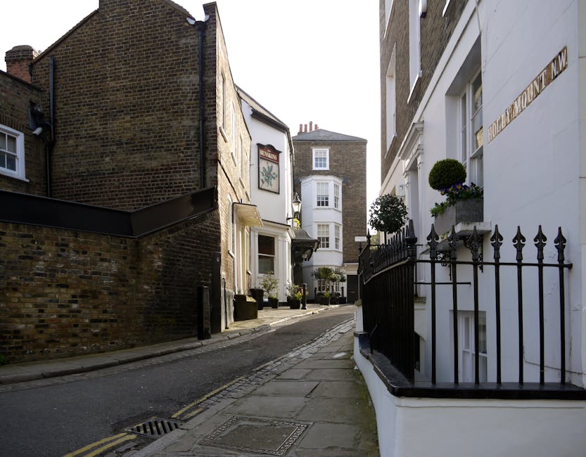A narrow, slightly inclined street scene in a residential area with a mix of brick and stucco buildings. On the right, there is a white building with potted plants and decorative black wrought iron fencing along the front. The building has a small window with a flower pot on the sill. To the left, there is a dark brick building with a small, white-framed window and a sign with a floral emblem on the façade. The pavement is made of concrete slabs, with a drainage grate visible in the foreground, and the street surface is asphalt with a double yellow line along the curb. Bright, natural daylight illuminates the scene, highlighting the textures of the brickwork, stucco finish, and plants, typical of urban residential streets where private rubbish collection or bespoke waste removal services, such as those provided by rubbishremovalwesthampstead.org.uk, might occur in the context of on-site clearance or alternative waste handling methods.