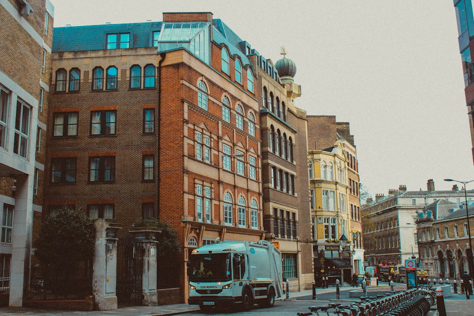 A street scene featuring a range of multi-story buildings, including one prominent red-brick structure with large, arched windows and decorative stonework, topped by a roof with multiple dormer windows and a small dome with a gold weather vane. To the right, a cream-colored building with ornate architectural details and bay windows is visible, along with other historic and modern buildings lining the street. In the foreground, a grey waste collection truck with a compact, boxy design and a white cab is parked near the sidewalk, which is lined with metal bike racks, bollards, and a sign indicating parking restrictions. The street appears busy with parked cars and people walking, set against a background of cloudy, overcast sky. The scene suggests an urban environment suitable for private waste disposal or rubbish removal services, reflecting local borough collection or alternative waste handling options.