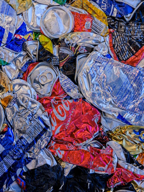 A close-up view of a pile of crushed and crumpled aluminium cans and plastic beverage bottles scattered on a flat surface. The cans feature various colours and brand labels, including a prominent red Coca-Cola can, with visible metallic surfaces, some with pull-tab openings, while others are flattened or dented. The plastic bottles are partially visible, with clear, transparent textures and some with crumpled labels. The background shows a mix of tangled packaging materials, capturing a chaotic assortment typical of waste collected for rubbish removal services, reflecting efforts towards waste collection or private disposal handling. The lighting appears natural, highlighting the reflective surfaces of the cans and the translucency of plastic bottles, emphasizing the textures and used condition of the waste items, which relate to the context of alternative waste management options outside of municipal collection systems.