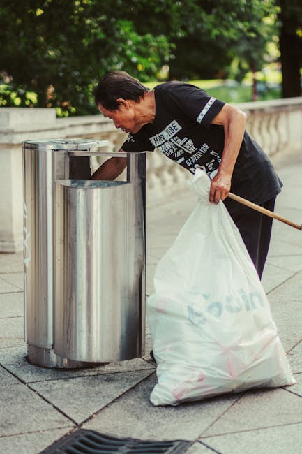 A woman with short dark hair, wearing a black t-shirt with white text and dark pants, is seen bending over to dispose of rubbish into a stainless steel public trash bin situated on a paved sidewalk. She is holding a large white plastic bag filled with waste in her left hand, while her right hand is reaching into the bin. The bin has a circular opening and a smooth, reflective surface, positioned next to a stone balustrade with decorative railings. In the background, there is lush greenery with trees and bushes, indicating an outdoor setting. The scene is illuminated by natural daylight, creating a clear and detailed view of the person and their activity related to rubbish disposal, which aligns with the context of private waste handling and waste removal services.