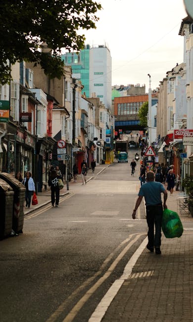 A busy street scene on a city street, with a mix of small retail shops and residential buildings on either side. In the foreground, a man in a blue shirt walks away from the camera, carrying a large green plastic bag filled with rubbish. Several pedestrians are walking along the pavement, some carrying shopping bags or talking. The buildings are a combination of brick and plaster facades, with some storefronts having awnings and signage, indicating local businesses. The street surface is paved with asphalt, featuring white double lines along the curb. In the background, taller modern office buildings with glass and steel exteriors, including a distinctive turquoise building, tower over the smaller structures. Trees are visible on the left side, providing some greenery amidst the urban environment. Overall, the scene depicts typical urban street activity, with occasional waste collection or disposal actions, such as the man carrying rubbish, which relates to independent rubbish removal services in the local area.
