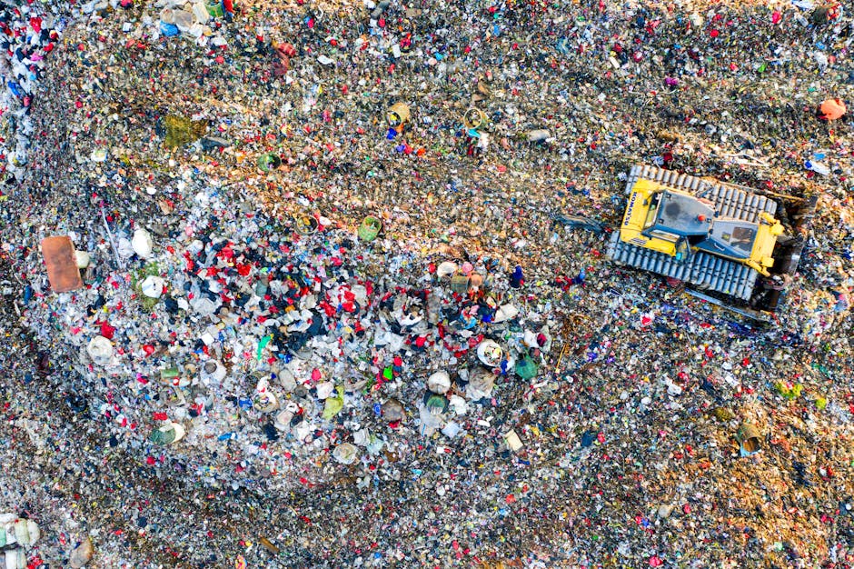 An aerial view of a large landfill site densely filled with mixed waste, including plastic bottles, containers, paper, and other refuse, forming a colorful and textured surface. The waste is spread unevenly across the site, with some heaps more concentrated and others more dispersed, creating a patchwork of different materials and colors. In the upper right area of the image, a yellow bulldozer with a metal blade attachment is visible, actively moving or leveling the waste. The environment is an open, outdoor terrain with natural sunlight casting minimal shadows, highlighting the variety of materials and their textures, from shiny plastics to duller paper and organic debris. The scene reflects the scale and complexity of waste management and disposal, illustrating the importance of rubbish removal services that handle such large quantities of refuse efficiently.