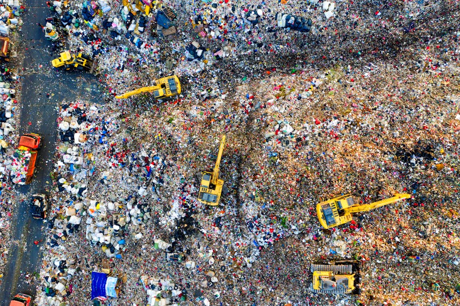 An aerial view of a large landfill site filled with a mixture of plastic bottles, packaging materials, and various types of waste debris. Several yellow excavators are actively sorting and managing the piles of refuse, with some of them digging into the waste to facilitate compaction or removal. The waste appears dense and unorganized, with different colors and textures of plastics, paper, and other materials visible throughout. Part of the site shows a dirt and gravel section where waste has been moved or deposited, with distinct piles and pathways for machinery access. In the background, a section of dark tarmac or paved area is visible, possibly part of a waste processing or collection vehicle route. The scene reflects a typical operation involving heavy machinery for waste handling and disposal, aligned with private or alternative rubbish removal services, such as those provided by companies like rubbishremovalwesthampstead.org.uk, highlighting the scale of waste management efforts outside municipal collection systems.