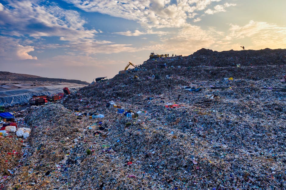 A large landfill site with multiple mounds of mixed waste materials extending across the landscape, under a sky filled with scattered clouds illuminated by a low sun, creating a warm glow. The mounds consist of various debris including plastics, textiles, and broken household items, with textured layers showing different colors such as blue, red, and black plastic fragments, alongside shredded paper and other refuse. Excavators and machinery are visible near the top of one mound, indicating ongoing waste handling processes. Tents or temporary coverings are pitched on the left side of the site, suggesting areas for sorting or storage. The environment appears industrial and expansive, emphasizing the scope of waste accumulation, relevant to discussions around alternative waste disposal methods like private rubbish removal services. The scene's composition highlights the scale and environmental impact of unmanaged waste, aligning with topics about independent collection and waste management options in the UK.